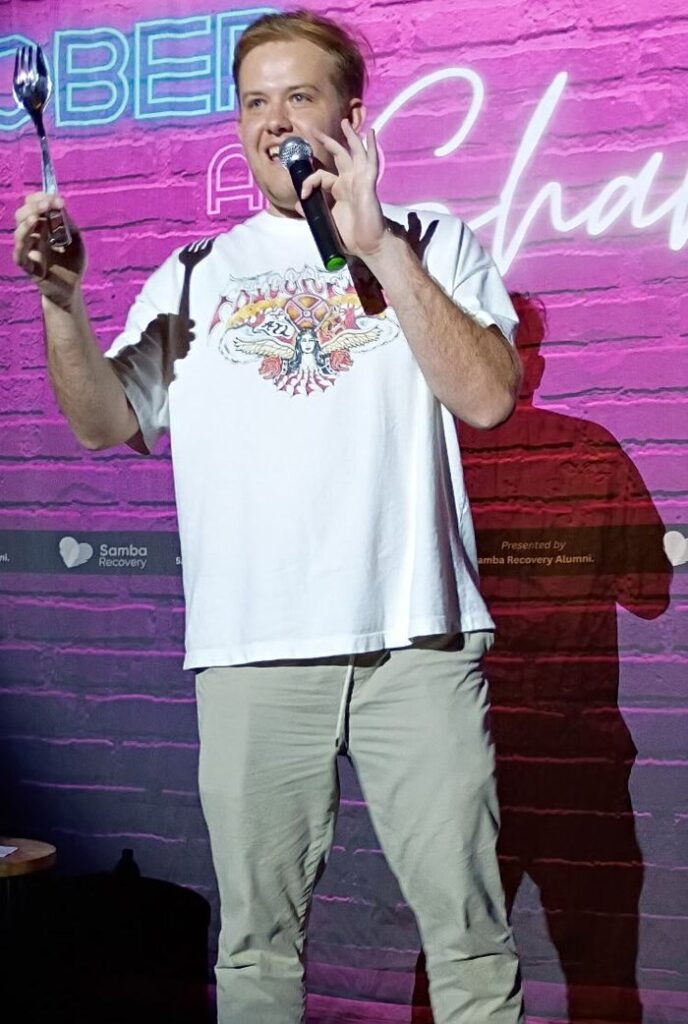 A stand-up comedian holding a microphone and a prop on the Sober and Shameless stage